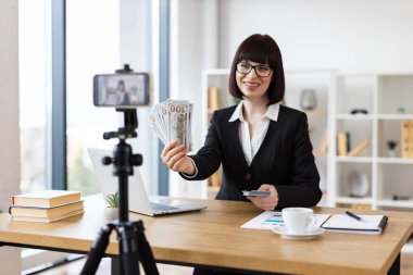 Ofisteki beyaz kadın videoda finansal konseptleri sunuyor. Nakit para tutarken gelir kaynakları, yatırımlar ve etkili para yönetimi tartışmaları.