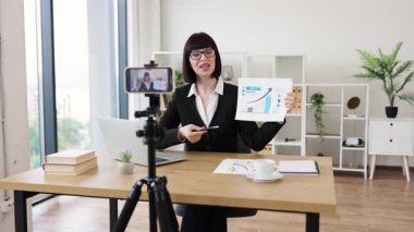 Beyaz kadın masa başında oturmuş iş performans istatistikleri ve büyüme analizleri hakkında blog videosu kaydediyor..