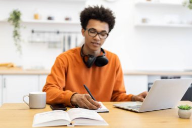 Gözlüklü genç bir adam evdeki dizüstü bilgisayarda çalışırken bir fincan kahve ve masada bir bitkiyle bir defter yazar..