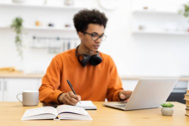 Gözlüklü genç bir adam masada çalışıyor, dizüstü bilgisayar kullanıyor ve notlar alıyor..