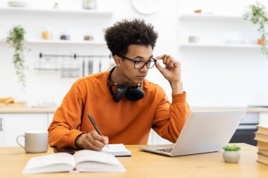 Gözlüklü genç bir adam masada çalışıyor, dizüstü bilgisayar kullanıyor ve notlar alıyor..