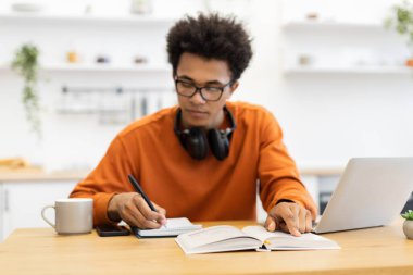 Gözlüklü genç bir adam, dizüstü bilgisayarı, kitabı ve defteri olan bir masada ders çalışıyor..