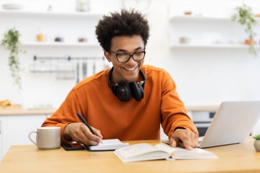 Gözlüklü genç bir adam ahşap bir masada çalışırken gülümsüyor, dizüstü bilgisayarı, kitabı ve defteriyle..