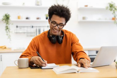 Genç siyah bir adam bir masada çalışırken dizüstü bilgisayarı, defteri ve kitabı ile gülümsüyor..