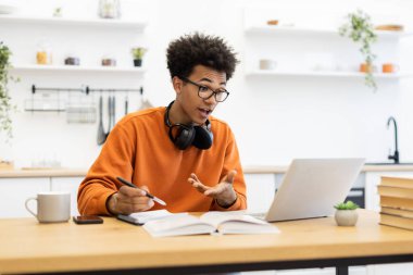 Gözlüklü genç bir adam video görüşmesi yapıyor. Parlak bir mutfaktaki ahşap bir masada el kol hareketi yapıyor ve notlar alıyor..