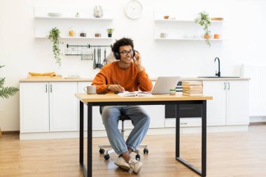 Kulaklıklı genç bir adam mutfaktaki bir masada dizüstü bilgisayarla çalışırken gülümsüyor..