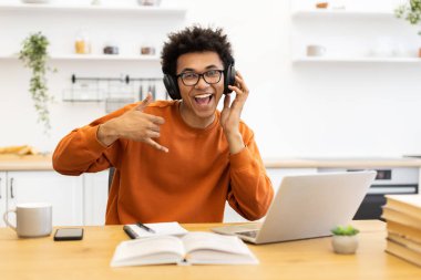 Kulaklık ve gözlük takan neşeli genç bir adam el hareketi yaparken gülümsüyor, evde dizüstü bilgisayarla çalışıyor..