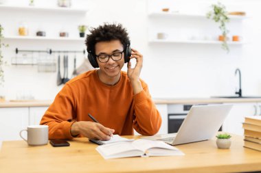 Kulaklık takan ve gözlük takan genç bir adam bir masada dizüstü bilgisayar ve bir fincan kahveyle notlar alırken gülümsüyor..