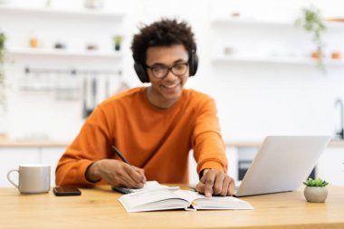 Kulaklık takan genç bir adam dizüstü bilgisayarı ve not defteriyle masada çalışırken gülümsüyor..