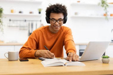 Kulaklık takan genç bir adam, dizüstü bilgisayarı, defteri ve kahve fincanıyla masada çalışırken gülümsüyor..