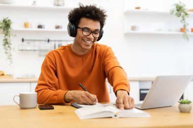 Gözlüklü ve kulaklıklı genç bir adam dizüstü bilgisayar ve defterle bir masada çalışırken gülümsüyor..