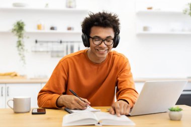 Kulaklık takan ve gözlük takan genç bir adam dizüstü bilgisayarla çalışırken gülümsüyor ve evdeki not defterine not alıyor..