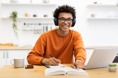 Gözlüklü ve kulaklıklı genç bir adam dizüstü bilgisayar ve defterle bir masada çalışırken gülümsüyor..
