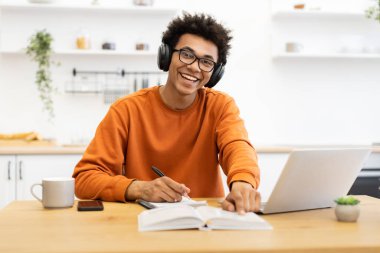 Kulaklık ve gözlüklü neşeli genç bir adam ahşap bir masada dizüstü bilgisayar, defter ve kahve fincanı ile çalışırken gülümsüyor..
