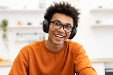 Gözlüklü ve kulaklıklı genç bir adam kameraya parlak bir şekilde gülümsüyor, turuncu bir kazak giyiyor..