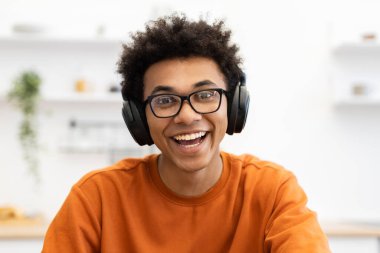 Gözlük ve kulaklık takan neşeli genç bir siyah adam kameraya bakarak geniş bir gülümseme sergiliyor..