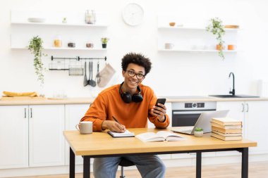 Gözlüklü genç bir adam dizüstü bilgisayarı üzerinde çalışırken gülümsüyor ve mutfakta notlar alıyor..