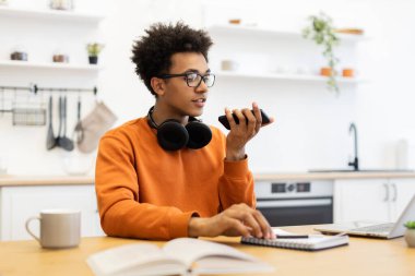 Gözlüklü genç bir adam, modern bir mutfak ortamında masa başında çalışırken telefonuna konuşuyor..