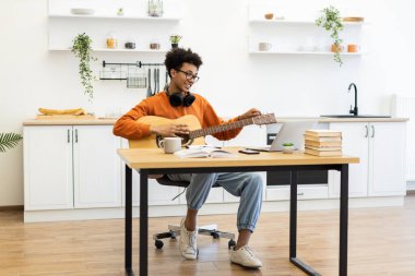 Gözlüklü genç bir adam mutfaktaki masada dizüstü bilgisayar kullanarak gitar akort ediyor..