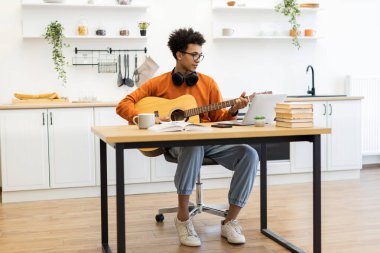 Gözlüklü genç bir adam modern bir mutfaktaki bir masada otururken akustik gitar çalar, yanında dizüstü bilgisayar ve kitaplarla..