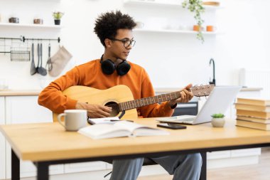 Gözlüklü genç bir adam parlak bir mutfak ortamında dizüstü bilgisayar kullanırken akustik gitar çalar..