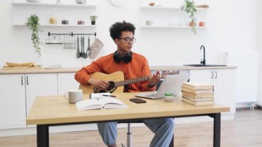 Gözlüklü genç bir adam elinde dizüstü bilgisayar ve defterle bir masada akustik gitar çalarken gülümsüyor..