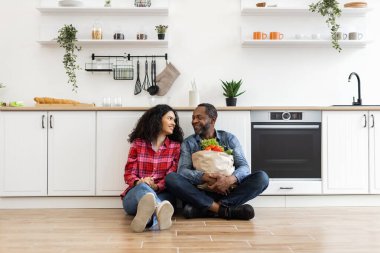 Gülümseyen bir çift mutfak zemininde oturup birbirlerine bakıyorlar. Adamın elinde bir torba erzak var..