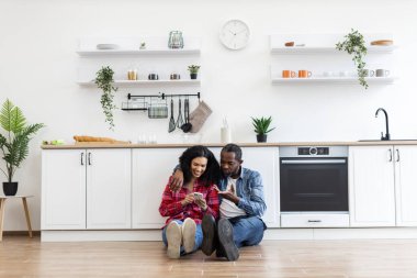 Gülümseyen bir çift, parlak, modern mutfaklarında yerde oturup birlikte telefona bakarlar..