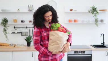 Kırmızı ekose gömlekli gülümseyen bir kadın mutfak ortamında içi taze yiyeceklerle dolu bir kese kağıdı tutuyor..