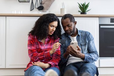 Bir çift mutfaklarında yerde oturmuş, bir kredi kartı ve akıllı telefona bakıyor..