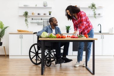 Farklı bir çift, parlak ve modern bir mutfakta, tekerlekli sandalye kullanan bir kişiyle birlikte yemek pişirmekten zevk alır..