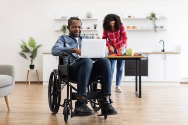 Tekerlekli sandalyedeki bir adam, ortağı mutfakta yemek hazırlarken dizüstü bilgisayar kullanır..