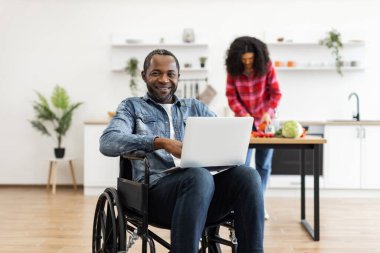 Tekerlekli sandalyedeki gülümseyen siyah bir adam modern bir mutfakta dizüstü bilgisayar kullanırken partneri arka planda yemek hazırlıyor..