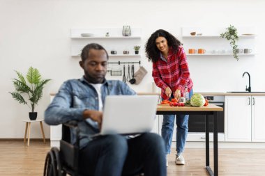 Bir kadın, ortağı mutfakta dizüstü bilgisayarla çalışırken yemek hazırlar. Adam tekerlekli sandalyede..