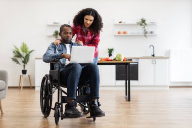 Tekerlekli sandalyedeki siyah bir adam ve bir kadın modern bir mutfak ortamında dizüstü bilgisayara bakıyorlar..