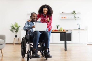 Tekerlekli sandalyedeki gülümseyen bir çift modern bir mutfak ortamında dizüstü bilgisayara bakıyorlar..