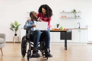 Tekerlekli sandalyedeki gülümseyen bir çift modern bir mutfak ortamında dizüstü bilgisayara bakıyorlar..