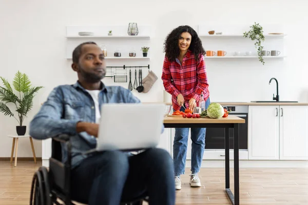 Tekerlekli sandalyedeki bir adam laptopta çalışırken partneri modern bir mutfakta yemek hazırlıyor..