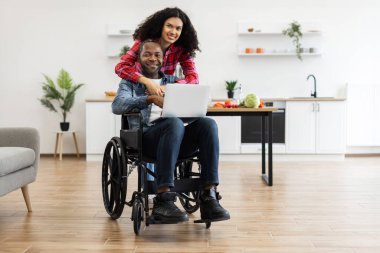 Gülümseyen bir çift, biri tekerlekli sandalyede dizüstü bilgisayar kullanıyor, diğeri de onu modern bir mutfak ortamında kucaklıyor..