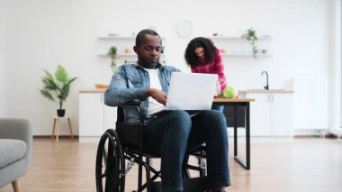 Tekerlekli sandalyedeki bir adam laptopta çalışırken ortağı mutfakta yemek hazırlıyor..