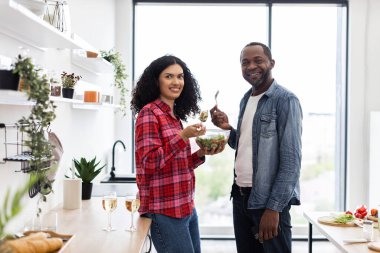 Gülümseyen bir çift, modern mutfak ortamında sağlıklı bir salatanın tadını çıkarır..