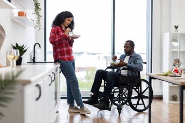 Çeşitli bir çift, modern bir mutfak ortamında, tekerlekli sandalye kullanan bir kişiyle birlikte sağlıklı bir yemek yiyor..