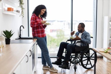 Değişik bir çift, parlak ve modern bir mutfakta sağlıklı bir salatanın tadını çıkarıyor. Bir kişi tekerlekli sandalyede..