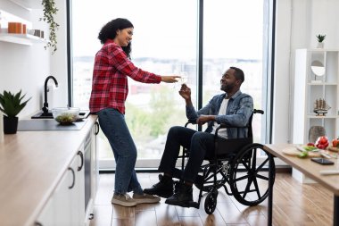 Bir kadın, modern mutfak ortamında tekerlekli sandalyedeki bir adama bir bardak şarap ikram ediyor. Gülümsüyorlar ve kadeh kaldırmak üzereler.