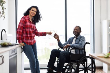 Modern bir mutfakta gülümseyen bir çift bir kadeh şarapla kadeh kaldırıyor, bir kişi tekerlekli sandalyede..