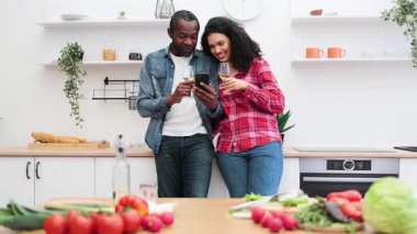 Melez bir çift, yeni ürünlerle çevrili modern bir mutfak ortamında telefona bakarken şaraptan zevk alır..