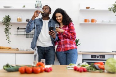 Bir çift mutfaklarında şarabın tadını çıkarırken telefona bakıyor. Taze sebzeler tezgahta..