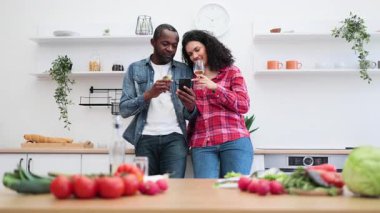 Bir çift, modern bir mutfakta, tezgahında taze sebzeler olan bir telefona bakarken şaraptan zevk alıyor..