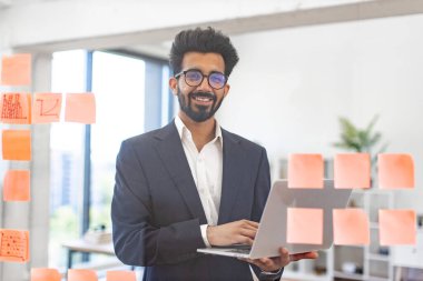 Hintli bir işadamı modern ofis ortamında dizüstü bilgisayarını tutarken gülümsüyor