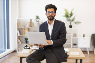 Takım elbiseli Hintli bir işadamı, bir ofis ortamında masa başında otururken dizüstü bilgisayarı üzerinde çalışıyor.